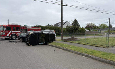 Black SUV resting on its side after a crash, with a red fire truck and firefighters at the scene in a residential street setting.