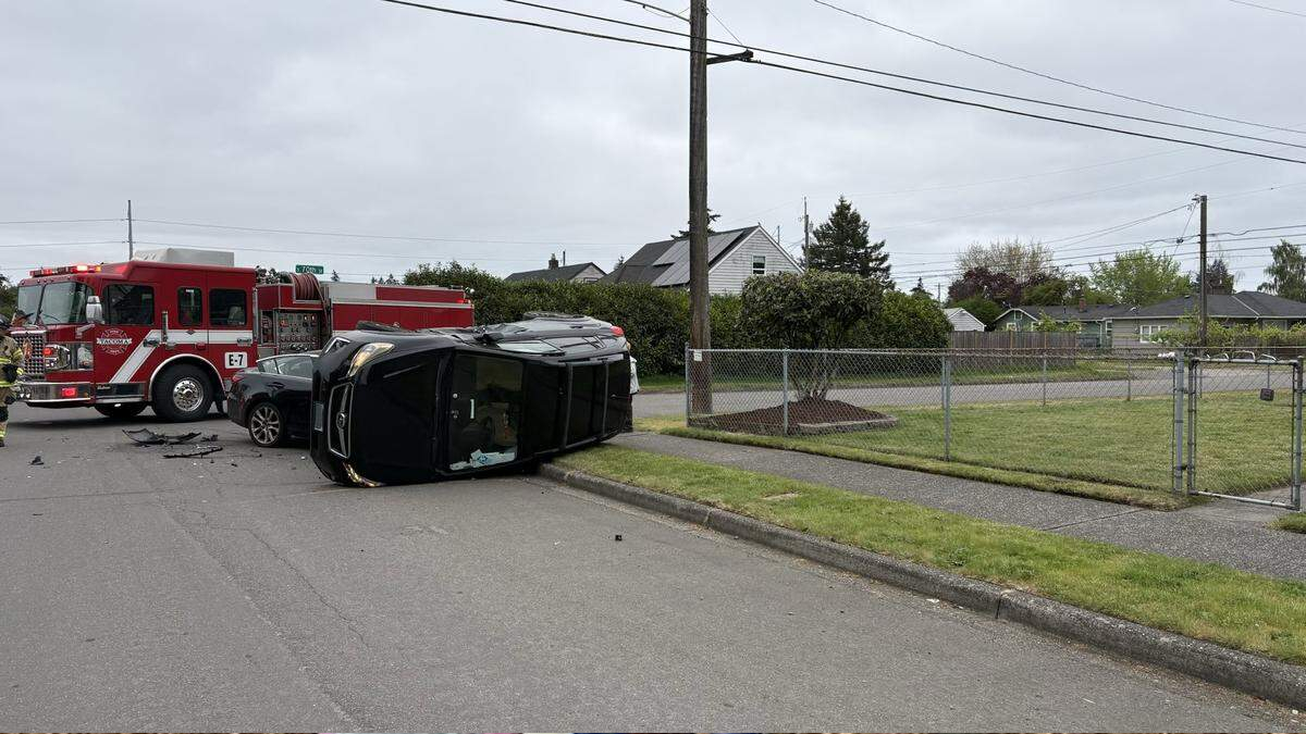 Black SUV resting on its side after a crash, with a red fire truck and firefighters at the scene in a residential street setting.