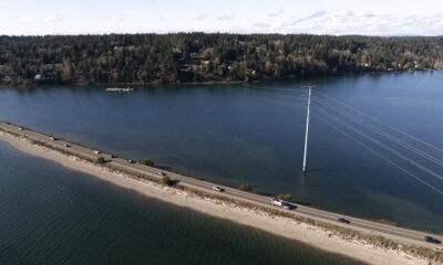 Aerial view of a road running along a flooded shoreline, with a tall power pole and lines arcing across deep water and a tree-lined bank in the distance.