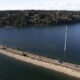 Aerial view of a road running along a flooded shoreline, with a tall power pole and lines arcing across deep water and a tree-lined bank in the distance.