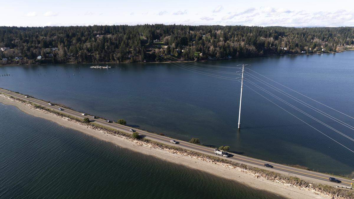 Aerial view of a road running along a flooded shoreline, with a tall power pole and lines arcing across deep water and a tree-lined bank in the distance.
