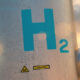 Industrial metal vessel with blue 'H2' label and caution EX signs in a sunny plant setting