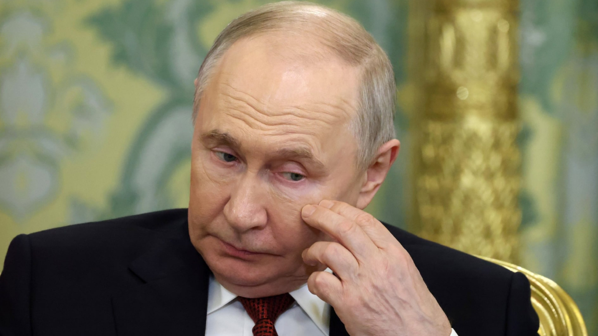 Older man in a dark suit rubs his eye, sitting against a gold-and-green backdrop with a contemplative expression.