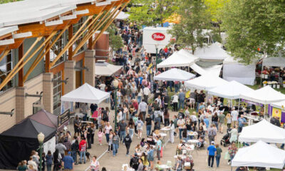 Busy outdoor market with white tents and stalls along a pedestrian path beside a brick building, people browsing and socializing.