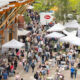 Busy outdoor market with white tents and stalls along a pedestrian path beside a brick building, people browsing and socializing.