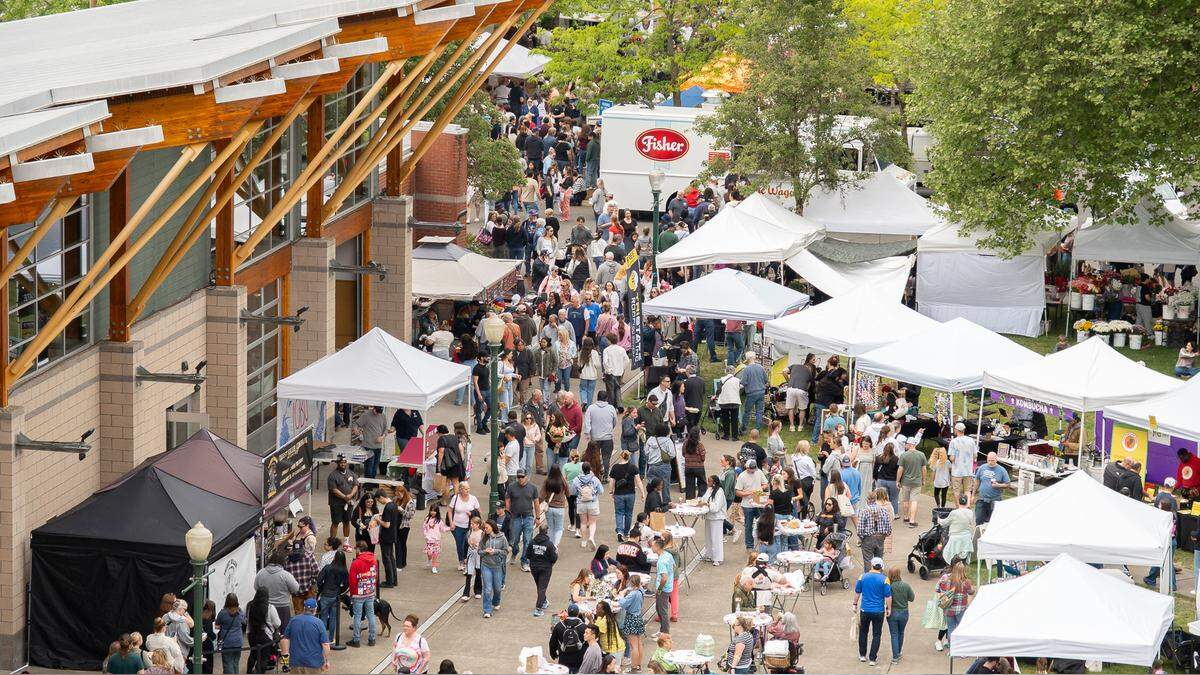 Busy outdoor market with white tents and stalls along a pedestrian path beside a brick building, people browsing and socializing.