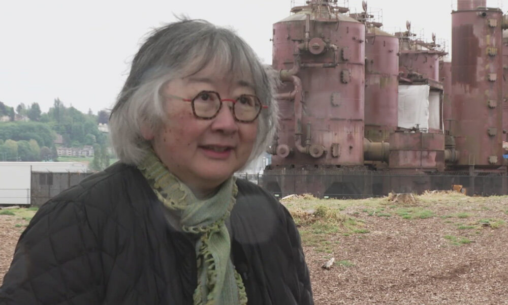 Older woman with gray hair and red glasses wearing a black quilted jacket and green scarf, outdoors near large rusted industrial machinery behind her.