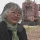Older woman with gray hair and red glasses wearing a black quilted jacket and green scarf, outdoors near large rusted industrial machinery behind her.