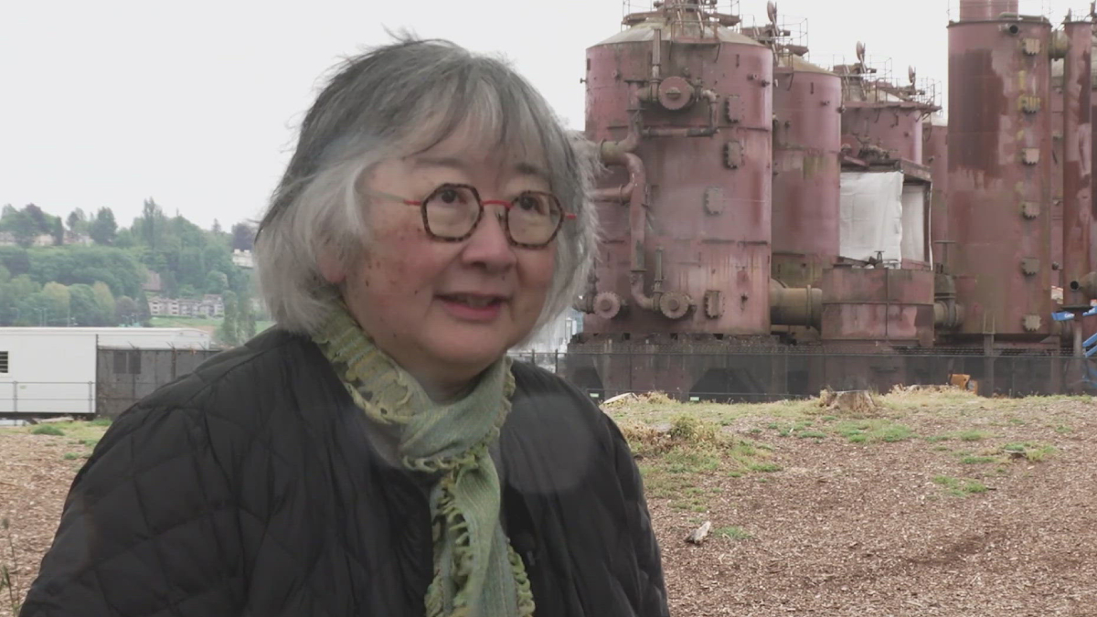 Older woman with gray hair and red glasses wearing a black quilted jacket and green scarf, outdoors near large rusted industrial machinery behind her.