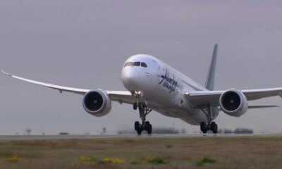 Alaska Airlines wide-body jet lifting off the runway, landing gear down, engines roaring against a cloudy sky.
