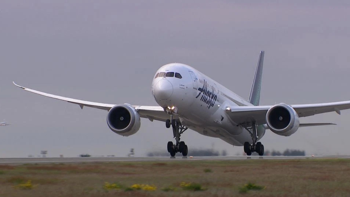 Alaska Airlines wide-body jet lifting off the runway, landing gear down, engines roaring against a cloudy sky.