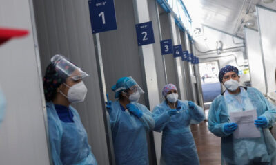 Healthcare workers in full PPE with masks and face shields standing in a corridor beside numbered bays 1–6.
