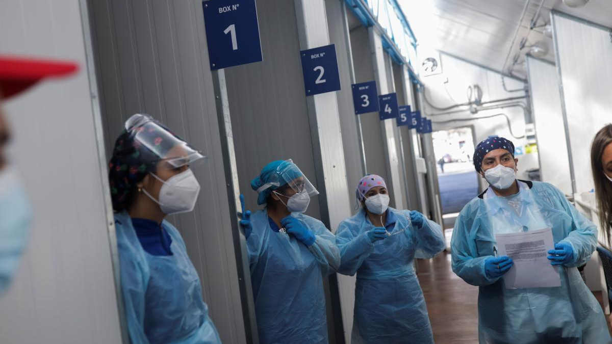 Healthcare workers in full PPE with masks and face shields standing in a corridor beside numbered bays 1–6.