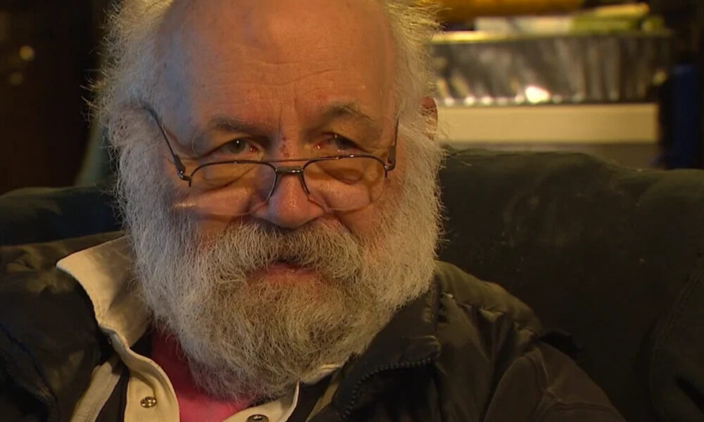 Close-up of an older man with a white beard and glasses, looking toward the camera while seated in a chair.