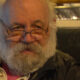 Close-up of an older man with a white beard and glasses, looking toward the camera while seated in a chair.