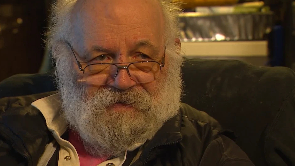 Close-up of an older man with a white beard and glasses, looking toward the camera while seated in a chair.