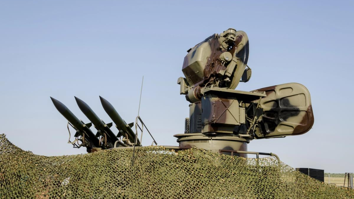 Military missile launcher mounted on an armored vehicle with four missile tubes, camouflaged with netting under a clear sky.