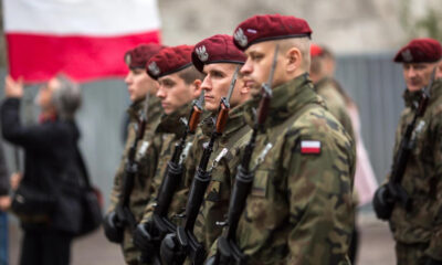 Polish soldiers in camouflage and red berets standing in formation, holding rifles during a ceremony with a Polish flag in the background.