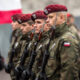 Polish soldiers in camouflage and red berets standing in formation, holding rifles during a ceremony with a Polish flag in the background.