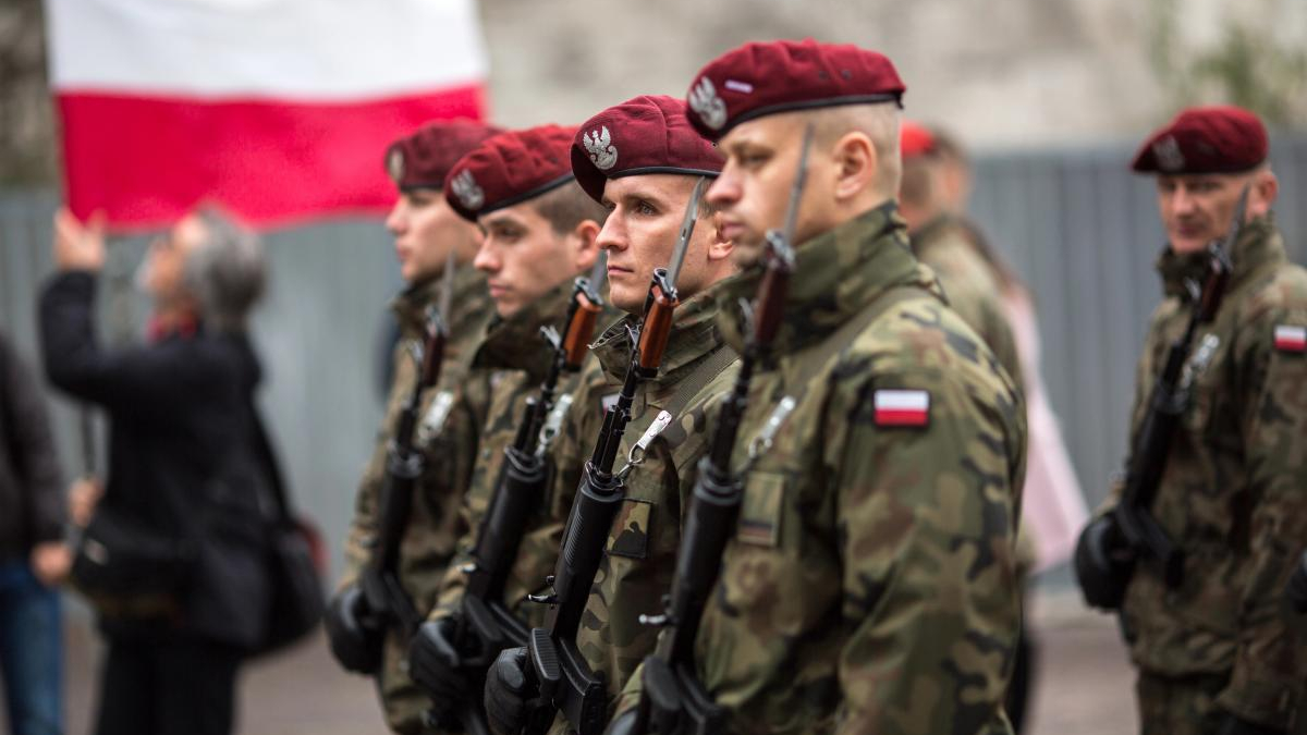 Polish soldiers in camouflage and red berets standing in formation, holding rifles during a ceremony with a Polish flag in the background.