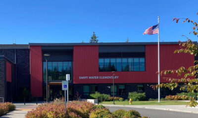 Front view of Swift Water Elementary School with red panels, large windows, and an American flag flying outside on a sunny day.