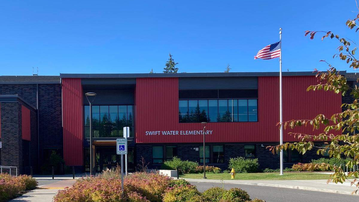 Front view of Swift Water Elementary School with red panels, large windows, and an American flag flying outside on a sunny day.