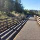 Paved pedestrian/cycle path flanked by wooden railings, sun casting long shadows, with trees and clear blue sky in a park-like setting.