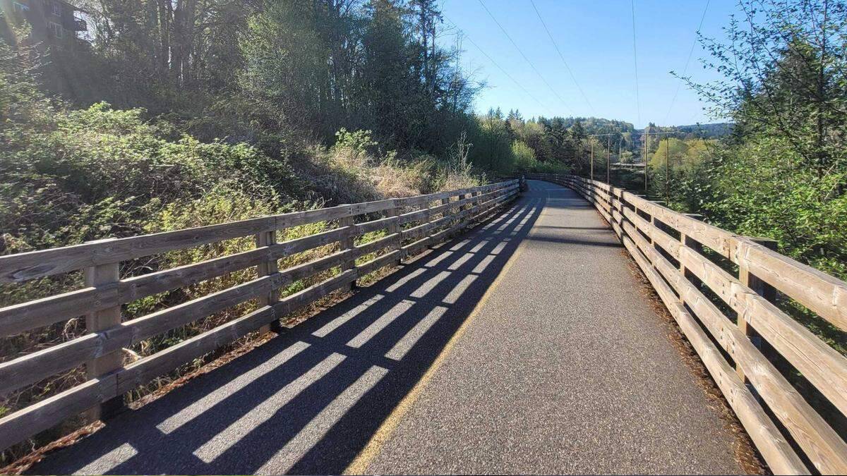 Paved pedestrian/cycle path flanked by wooden railings, sun casting long shadows, with trees and clear blue sky in a park-like setting.
