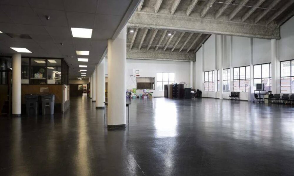Empty industrial-style hall with white columns, polished dark floor, and large windows along the right side; storage and desks near the left wall