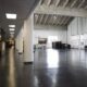 Empty industrial-style hall with white columns, polished dark floor, and large windows along the right side; storage and desks near the left wall