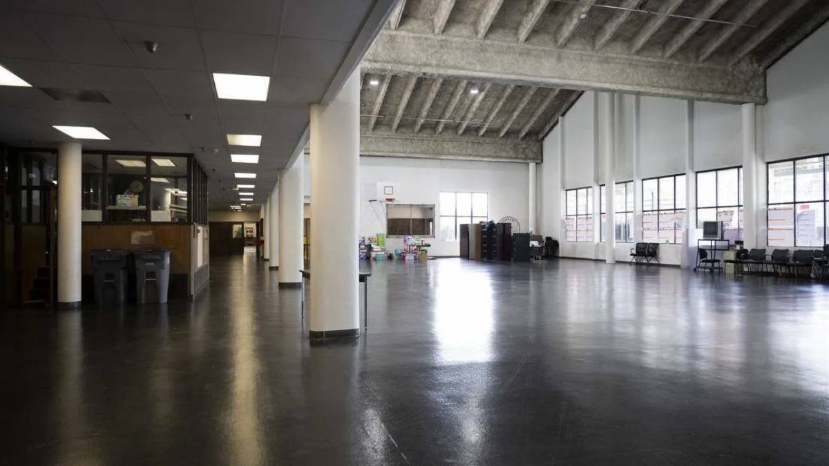 Empty industrial-style hall with white columns, polished dark floor, and large windows along the right side; storage and desks near the left wall