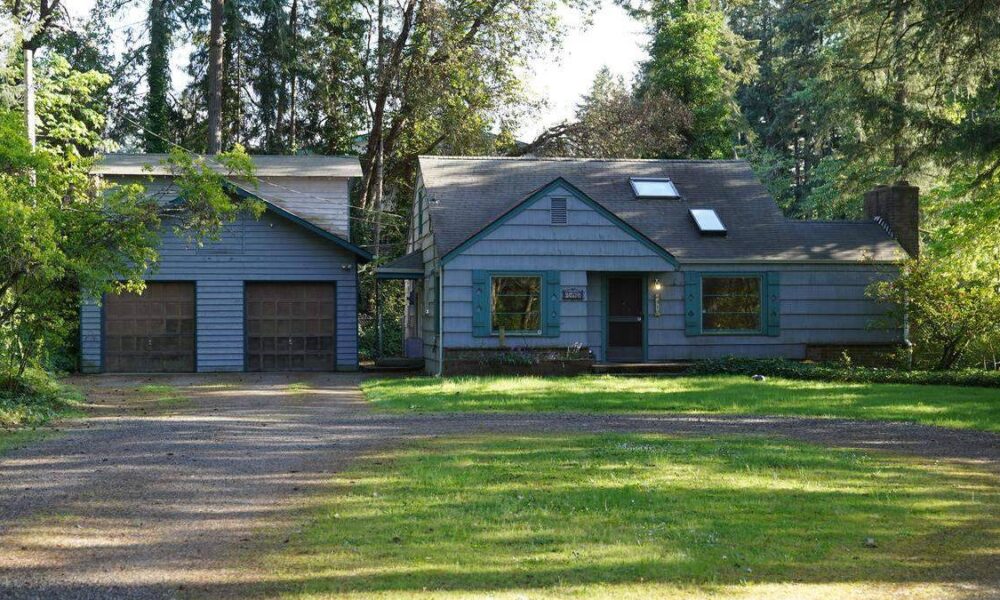 Blue ranch-style house with green trim and a two-car detached garage, set among trees and a gravel driveway on a sunny day.