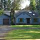 Blue ranch-style house with green trim and a two-car detached garage, set among trees and a gravel driveway on a sunny day.