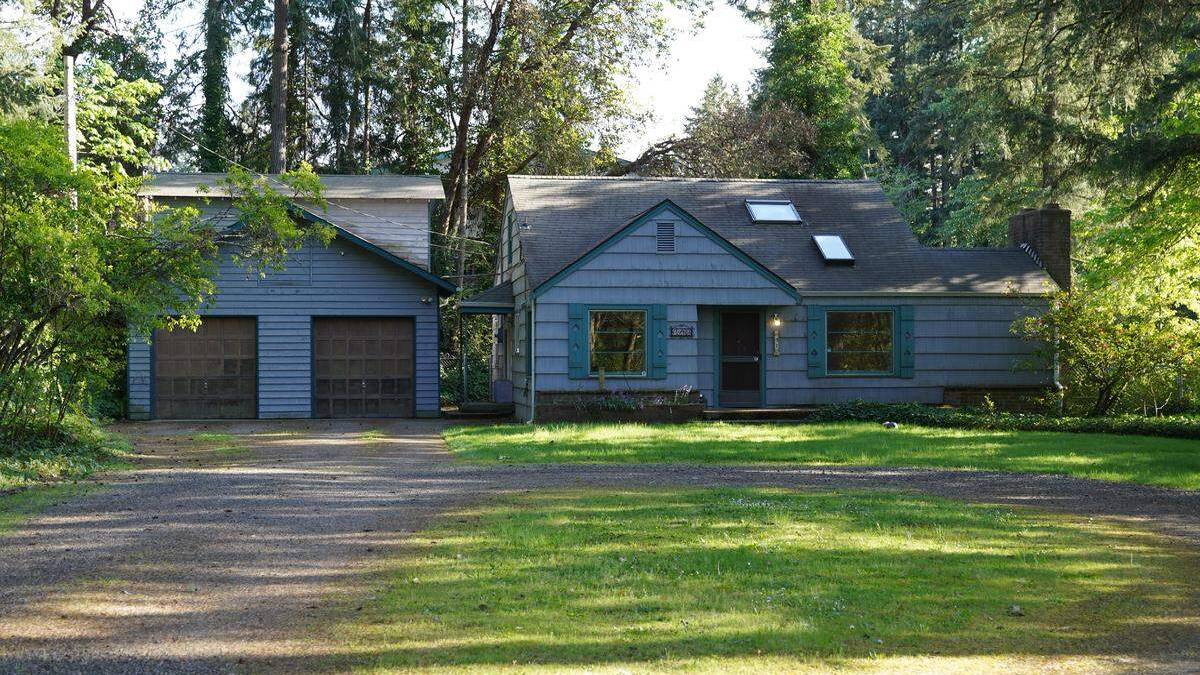 Blue ranch-style house with green trim and a two-car detached garage, set among trees and a gravel driveway on a sunny day.