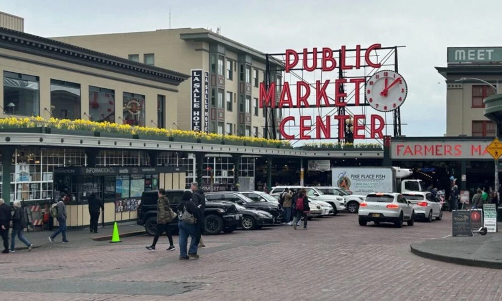 Public Market Center sign over Pike Place Market with pedestrians and parked cars on a brick-paved street.