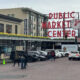 Public Market Center sign over Pike Place Market with pedestrians and parked cars on a brick-paved street.