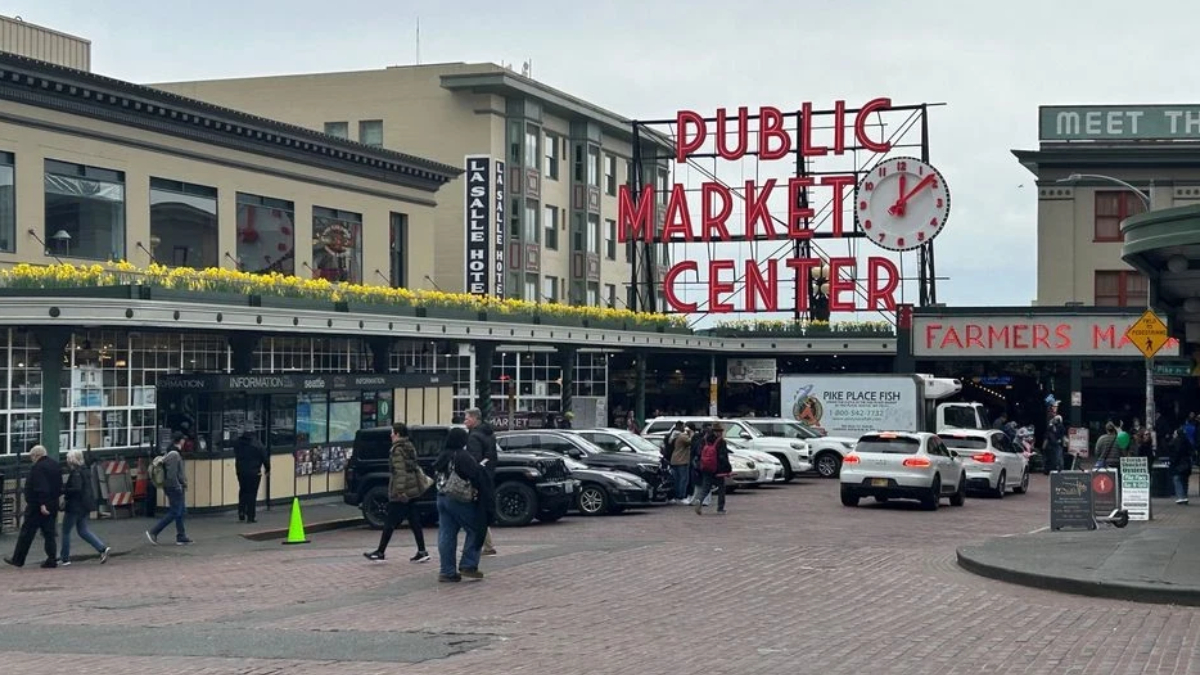 Public Market Center sign over Pike Place Market with pedestrians and parked cars on a brick-paved street.