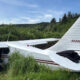 Small white single-engine plane crashed in tall grass; firefighter kneels at the open cockpit door by a damaged wing.