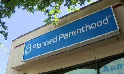 Blue storefront sign reading 'Planned Parenthood' on a building, with tree branches overhead.