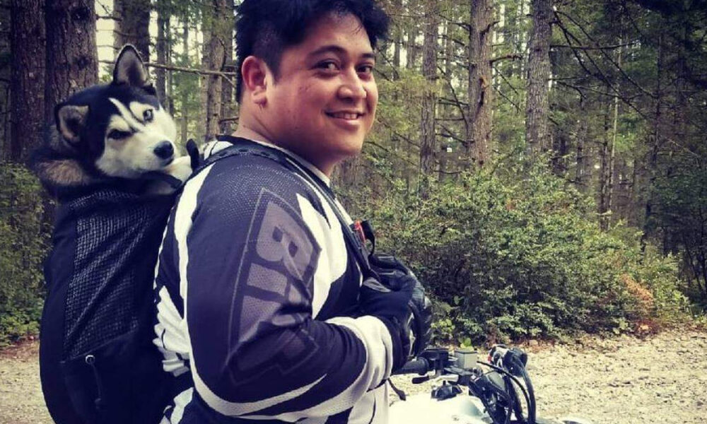 Smiling person wearing motorcycle gear in a forest, with a husky riding in a backpack-style dog carrier.