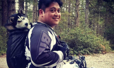 Smiling person wearing motorcycle gear in a forest, with a husky riding in a backpack-style dog carrier.