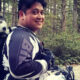 Smiling person wearing motorcycle gear in a forest, with a husky riding in a backpack-style dog carrier.