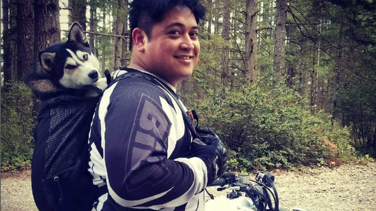 Smiling person wearing motorcycle gear in a forest, with a husky riding in a backpack-style dog carrier.