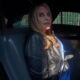 Woman with blonde hair in a black leather jacket sits in the back seat of a dark car, looking at the camera.