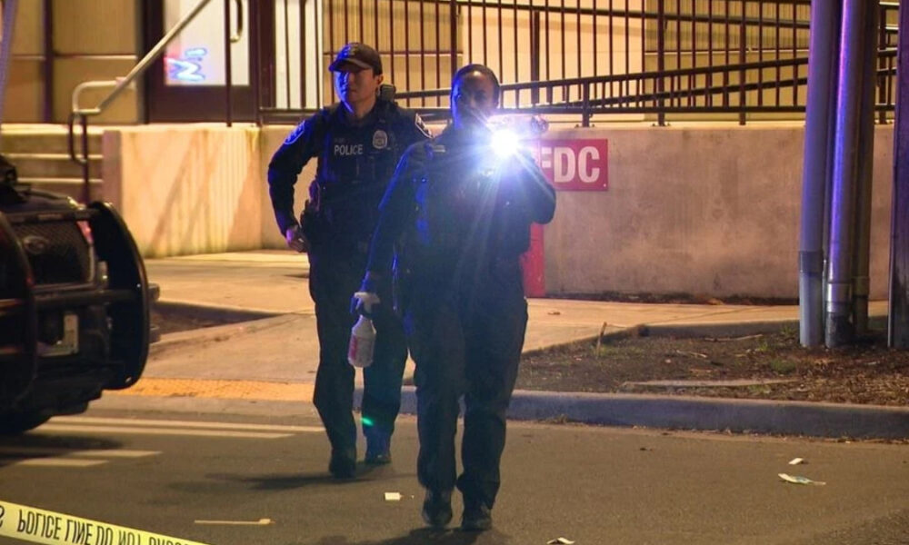 Two police officers walk toward the camera at a nighttime crime scene, one shining a bright flashlight toward the lens while others guard behind. Police tape borders the scene.