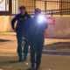 Two police officers walk toward the camera at a nighttime crime scene, one shining a bright flashlight toward the lens while others guard behind. Police tape borders the scene.
