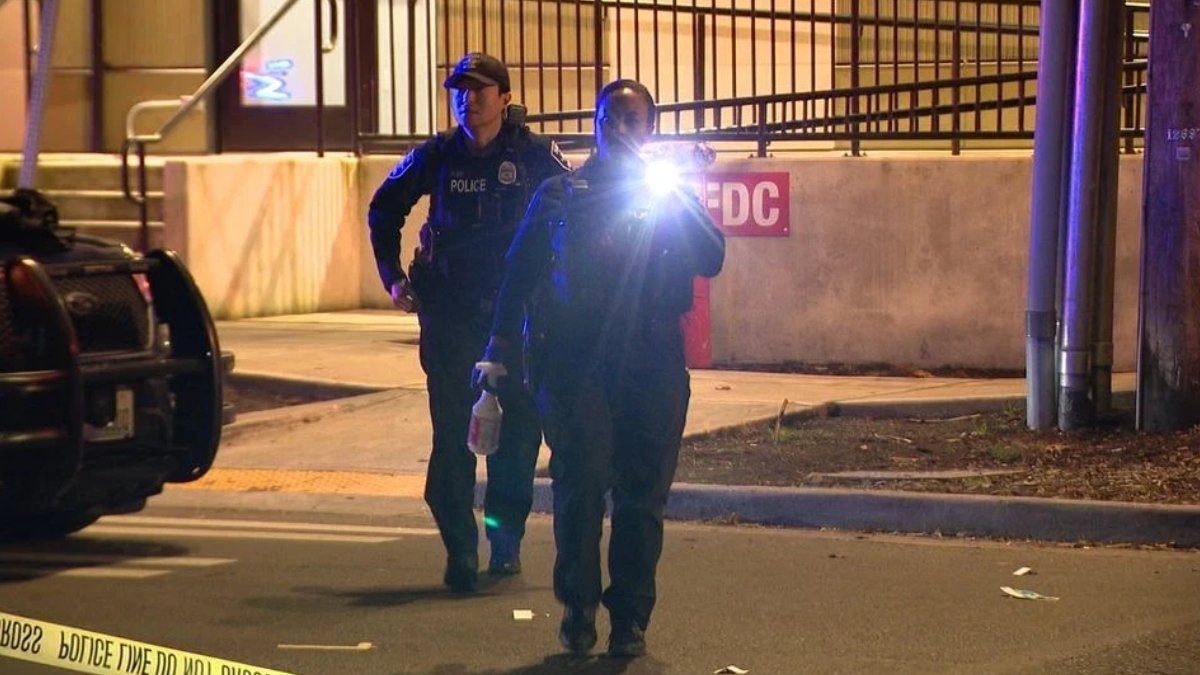 Two police officers walk toward the camera at a nighttime crime scene, one shining a bright flashlight toward the lens while others guard behind. Police tape borders the scene.