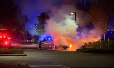 Nighttime scene of a car on fire in a parking lot, smoke billowing as firefighters battle the blaze with red and blue emergency lights nearby.