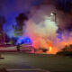 Nighttime scene of a car on fire in a parking lot, smoke billowing as firefighters battle the blaze with red and blue emergency lights nearby.