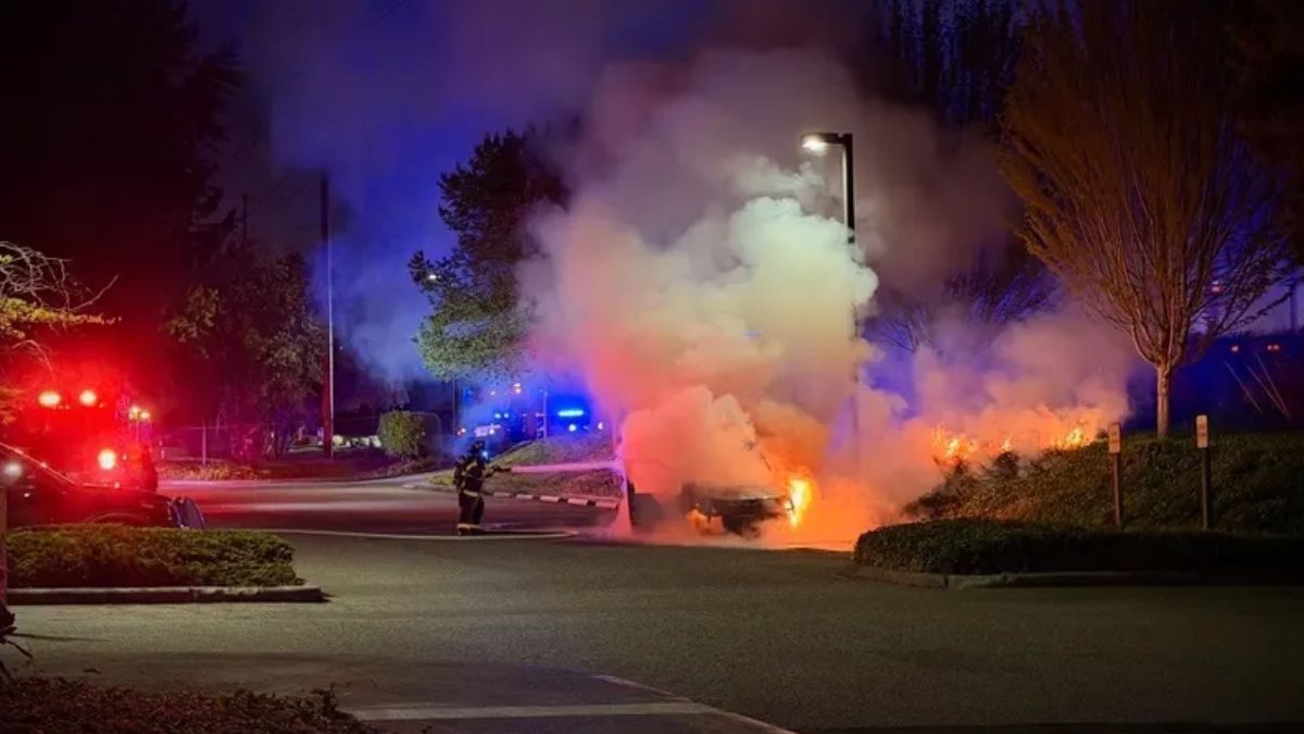 Nighttime scene of a car on fire in a parking lot, smoke billowing as firefighters battle the blaze with red and blue emergency lights nearby.